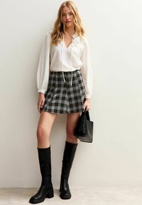 Woman wearing white long-sleeve blouse, gray plaid mini skirt, black knee-high boots, holding black handbag, standing against light wall.