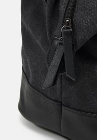 Sac à dos noir avec un corps en tissu texturé, un fond en cuir et une fermeture éclair noire. Dispose de tirettes à fermeture éclair noires plates et d'une forme structurée.
