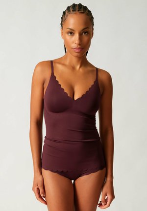 Frau mit geflochtenem Haar, die ein eng anliegendes dunkelviolettes Camisole und passende Unterwäsche mit gewellten Kanten trägt, stehend vor einem einfarbigen hellen Hintergrund.