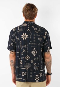 Chemise à manches courtes en tissu bleu marine, avec des motifs géométriques et abstraits crème. Coupe ample avec un col rabattu et un ourlet carré.