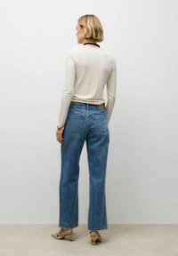 Vrouw die zich omgekeerd bevindt, gekleed in een crèmewitte langemouwen top, blauwe hoge taille rechte jeans, een zwarte kraal ketting, en slangenprint kitten heels.