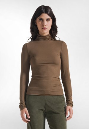 Femme aux longs cheveux foncés portant un pull à col roulé marron et un pantalon vert, debout devant un fond blanc uni.