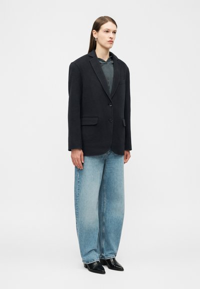 Ženska z dolgimi ravnimi lasmi nosi črn blazer čez temno jopa, široke modre kavbojke s širokimi hlačnicami in črne koničaste čevlje, stoji pred enobarvnim ozadjem.