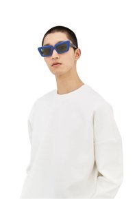 Occhiali da sole blu a forma quadrata con finitura opaca e lenti oscurate, indossati con un ampio sudario bianco semplice.