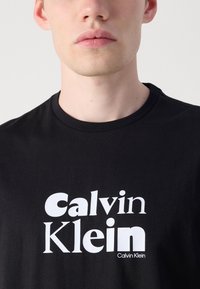 Juodas medvilnės marškinėliai su dideliu baltu logotipu "Calvin Klein" moderniu šriftu. Apvalus kaklas ir glotni tekstūra.