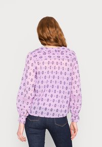Blusa lilás clara com estampado floral, mangas bufantes longas, detalhe de ombro franzido e textura subtil, combinada com jeans de denim escuro.