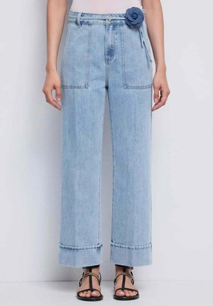 Jeans de mezclilla azul claro de tiro alto con bolsillos frontales de parche, dobladillos doblados y un detalle de cinturón con flores azules, usados con sandalias negras.