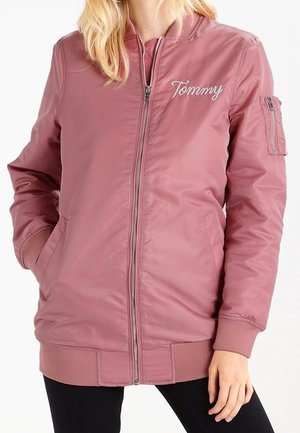 Kobieta nosząca bomberkę w kolorze pudrowego różu z zamkiem błyskawicznym, z wyhaftowanym napisem "Tommy" na piersi oraz kieszeniami z przodu i na rękawie.