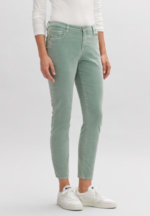 Personne portant un pantalon slim vert en velours côtelé, un haut blanc à manches longues et des baskets blanches, debout sur un fond uni.
