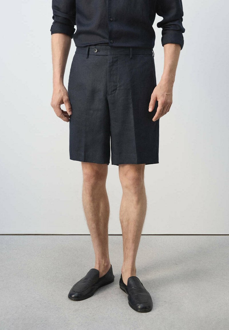 Homme portant un short sombre sur mesure, une chemise noire à manches longues retroussées et des chaussures noires sans lacets, debout sur un sol gris.