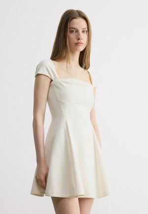 Jauna sieviete ar taisniem brūniem matiem baltā īsā kleitā ar piedurknēm, stāvot pret vienkāršu gaišu fonu.