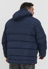 Marineblå dunjakke med hætte, der har kviltede sektioner. Materialet ser glat ud med en mat finish. Indeholder en lille læderakcent ved kanten.