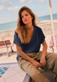 Un haut marine à col en V avec des manches courtes, associé à un pantalon beige taille haute, assis à une table carrelée près de l'océan.