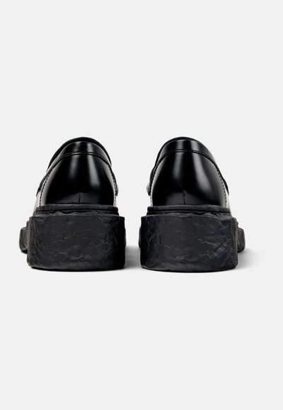 Schwarze Lederschuhe mit einer massiven, strukturierten schwarzen Sohle. Sie verfügen über eine runde Zehenpartie und eine gebogene Ferse, mit sichtbaren Nähten entlang des Obermaterials.