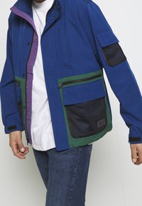 Blå flerfickad jacka med gröna och lila detaljer, med dragkedja och nätfickor, hög krage och justerbara ärmslut.