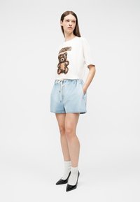 Γυναίκα που φοράει λευκό T-shirt Moschino με αρκουδάκι, ανοιχτό μπλε σόρτς με κορδόνια, λευκές κάλτσες και μαύρες μυτερές γόβες, στέκεται με τα χέρια στις τσέπες.