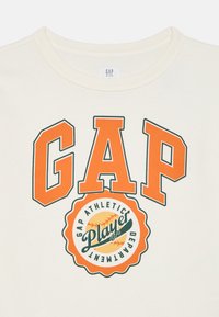 Krémový bavlnený sweatshir s výrazným nápisom "GAP" v oranžovej farbe, pod ktorým je okrúhly grafický prvok so slovami "GAP ATHLETIC PLAYERS DEPARTMENT."