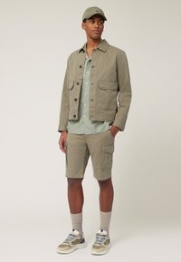 Licht olijfgroene katoen jack met vier voorzakken, gecombineerd met een overhemd met knopen en een korte broek. Draag het met sneakers en een bijpassende pet.