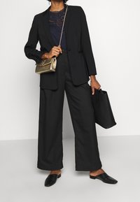 Černý přizpůsobený blazer přes námořnické tričko, spojený se širokými černými kalhotami. Doplňky zahrnují zlatou kabelku a černé texturované mokasíny.