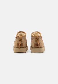 Brown suede ankle boots with shearling lining, flat rubber sole, and decorative leather accents. Features branded back label and tied detail.