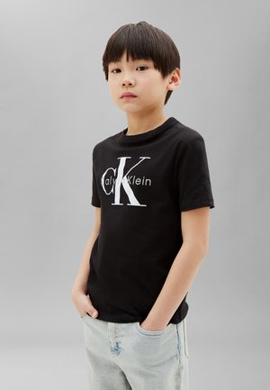 Garçon aux cheveux courts et foncés portant un t-shirt noir Calvin Klein et un jean bleu clair, debout les mains dans les poches devant un fond gris uni.