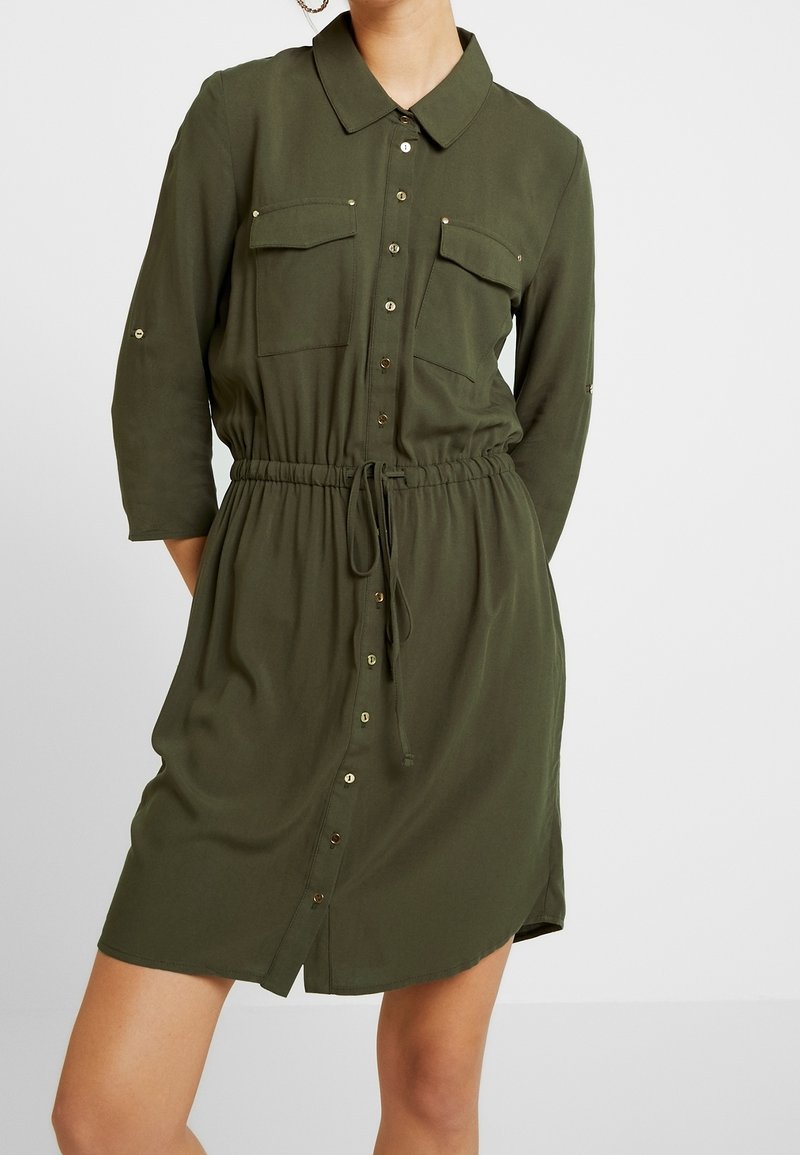 Femme portant une robe-chemise vert olive à boutons, manches longues, deux poches poitrine, et ceinture à cordon.