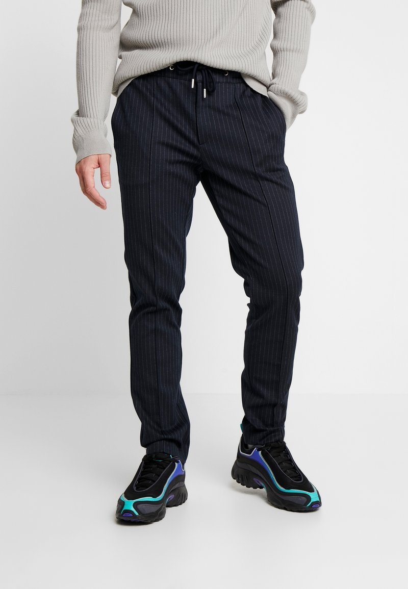 Homme portant un pantalon noir à fines rayures avec cordon de serrage et des baskets noires avec des accents bleus et turquoise, debout avec une main dans la poche.