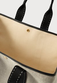 Μπεζ καμβάς τσάντα tote με μαύρους χειρολαβές, μαύρο περίγραμμα και μικρό χρυσό κουμπί κλείσιμο στο εσωτερικό της ανοιχτής κορυφής.