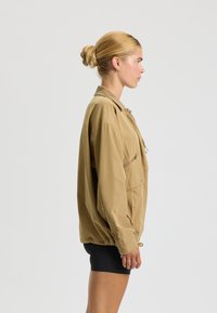 Khaki leichte Jacke mit einer Reißverschlusstasche, lockerer Passform, Stehkragen und gerafftem Saum. Glatte Textur mit minimalen Hardwaredetails.