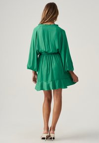 Kurzes grünes Kleid mit fließender Silhouette, gerafftem Taillenbund und Rüschenausschnitt; aus leichtem Stoff mit langen Ärmeln und ausgestelltem Saum.