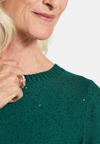 Grüner Strickpullover mit geripptem Ausschnitt, der ein strukturiertes Muster und verstreute glitzernde Akzente aufweist. Lange Ärmel, weiches Material.