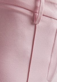 Ljust rosa tyg med en slät textur. Har en subtil sömdetalj och en ögla för bälte eller knäppning. Enkel, minimalistisk design.