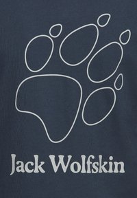Λευκό περίγραμμα αποτυπώματος πατούσας λύκου πάνω από το κείμενο "Jack Wolfskin" σε σκούρο μπλε υφασμάτινο φόντο.