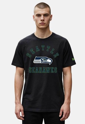 Junger Mann mit kurzen Haaren, der ein schwarzes Seattle Seahawks T-Shirt mit dem Teamlogo und Namen auf der Vorderseite trägt.