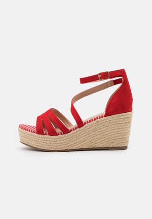 Sandales compensées en suède rouge avec une plateforme en jute tressé. Présente des brides croisées et une sangle à la cheville avec une boucle. Détail de semelle intérieure rayée.