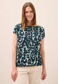 Mujer sonriente con cabello hasta los hombros, vistiendo una blusa de manga corta de patrón abstracto en tonos verde azulado y negro, y pantalones claros beige, frente a un fondo liso.