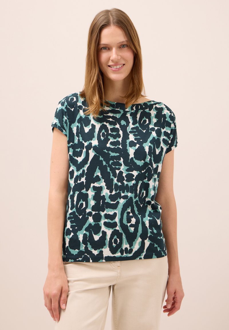 Mujer sonriente con cabello hasta los hombros, vistiendo una blusa de manga corta de patrón abstracto en tonos verde azulado y negro, y pantalones claros beige, frente a un fondo liso.