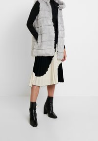 Chaleco de piel sintética gris con rayas horizontales, cierre de cremallera y capucha. Llevado sobre una blusa negra de manga larga y una falda plisada beige y negra, combinada con botines negros.