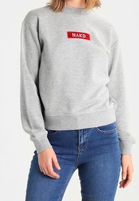 Sudadera gris de tejido suave con dobladillo acanalado, que presenta un parche rectangular rojo centrado en el pecho con la inscripción "NAKD".