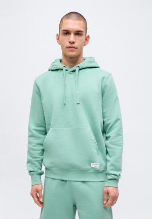 Νέος άνδρας που φοράει ανοιχτό πράσινο φούτερ με μπροστινή τσέπη και ταιριαστό παντελόνι φόρμας, στέκεται μπροστά σε απλό λευκό φόντο.