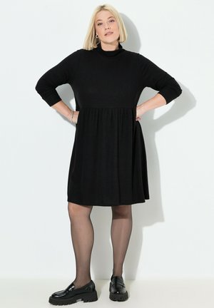 Schwarzes Kleid mit hohem Kragen, langen Ärmeln und tailliertem Oberteil; der ausgestellte Rock endet über dem Knie. Getragen mit schwarzen Slippern und Strumpfhosen.
