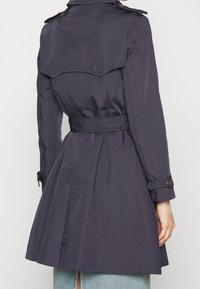 Marinblå trenchcoat med cinchad midja och plisserad baksidesdetalj. Har axelremmar och bältesmanschetter, tillverkad av ett slätt, hållbart tyg.