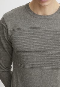 Uomo che indossa un maglione a girocollo con texture e motivi nelle tonalità di grigio, accorciato dal collo fino a metà torso, su uno sfondo semplice.
