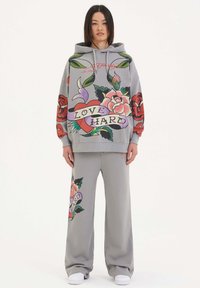 Grå huvtröja med blommiga motiv och texten "LOVE HARD," i kombination med grå vida byxor med liknande mönster. Vita sneakers.
