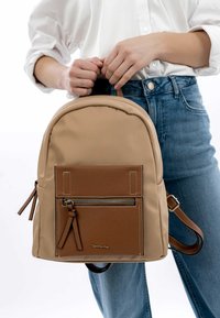 Sac à dos beige avec une finition lisse, poche texturée marron, fermeture à glissière en métal argenté et bretelles ajustables. Design minimaliste avec des coutures accentuées.