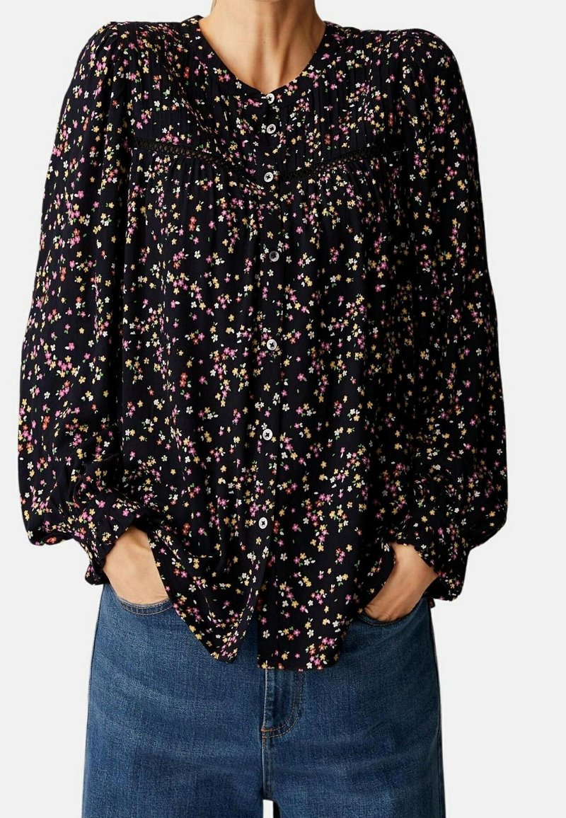 Femme portant un chemisier noir à fleurs boutonné avec de longues manches bouffantes et un jean bleu, mains dans les poches avant.