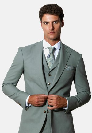 Man fastening buttons on light green three-piece suit with patterned green tie and pocket square against white background.