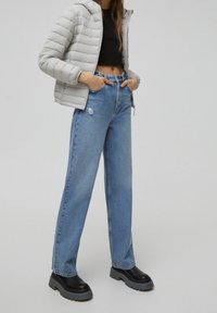 Femme portant une doudoune gris clair, un top court noir, un jean bleu taille haute avec une déchirure et des bottes noires épaisses, debout les mains dans les poches.