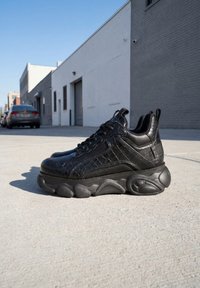 Zapatilla negra de estilo chunky con cuero texturizado y suela gruesa escultórica colocada en una acera urbana bajo un cielo despejado.