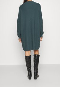 Abito a coste color teal con spalle scese, maniche lunghe e vestibilità comoda, abbinato a stivali neri lucidi fino al ginocchio.
